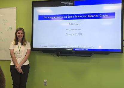 JCU mathematics major Reilly Lewis stands next to classroom display screen with title slide of her mathematics research presentation